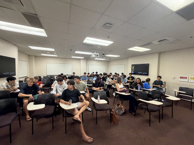 A large group of people seated in a classroom or conference room with rows of desks and chairs, watching something attentively.