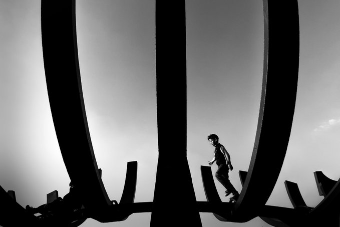A black and white silhouette photograph shows a boy walking on a metal structure against a bright sky.