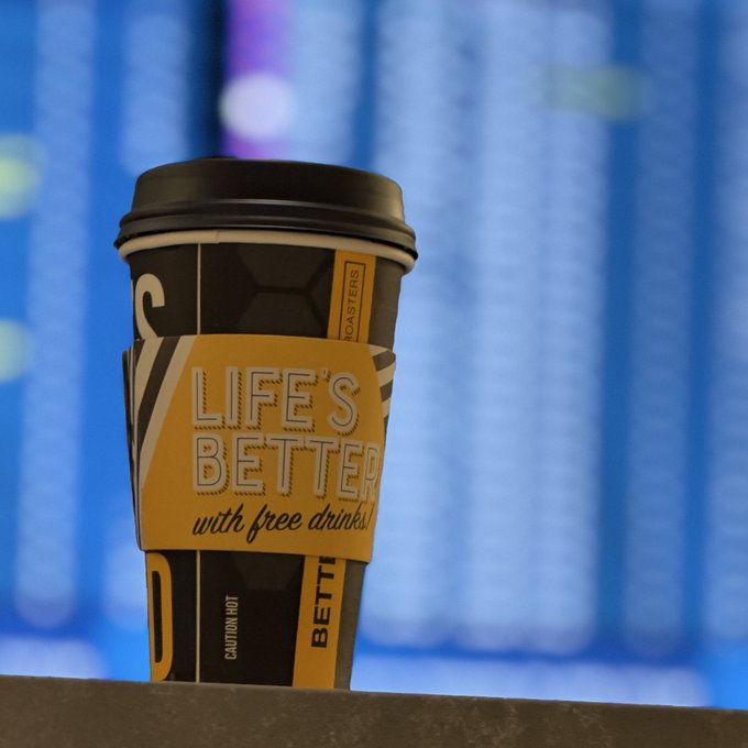 A close-up of a coffee cup with a black and yellow design featuring the text 'LIFE'S BETTER with free drinks' and 'BETTER ROASTERS', set against a blurred blue digital background.