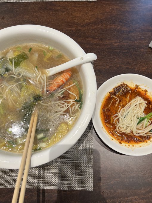 A delicious bowl of seafood noodle soup featuring a large shrimp, alongside a smaller dish of noodles with a spicy chili oil dipping sauce, with chopsticks ready for the meal.