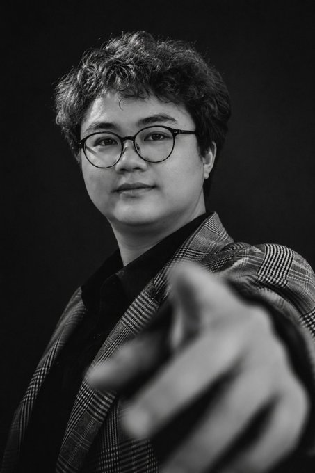 A black-and-white portrait of a young man with curly hair and round glasses wearing a plaid blazer, pointing toward the camera.
