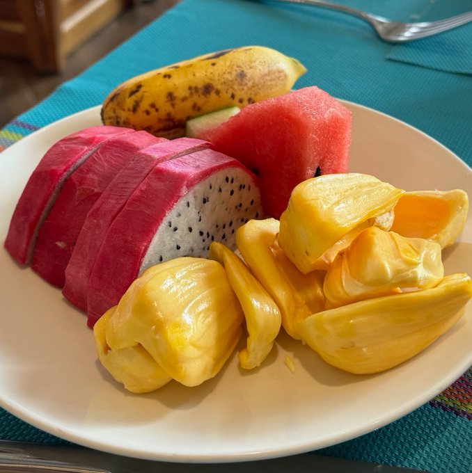 A white plate holds an assortment of fresh tropical fruit, including a sliced pink dragon fruit, yellow jackfruit pods, red watermelon, and a small banana, set on a teal placemat.