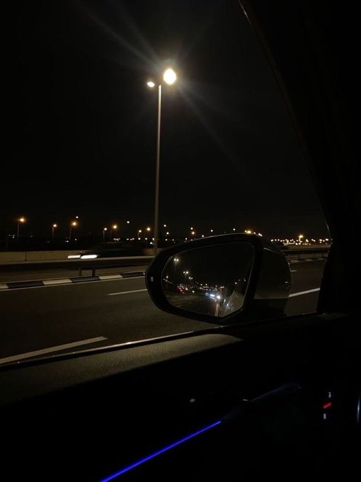 A nighttime drive captured from inside a car, with the side mirror reflecting headlights from vehicles behind on a highway illuminated by streetlights. The dark, atmospheric scene ties to the caption's mention of "night rides," evoking the quiet anticipation of an arrival tomorrow.