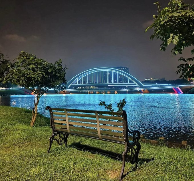 An empty park bench on grassy ground faces a glowing arched bridge illuminated in bright blue over a calm river at night, offering a quiet reflective setting that pairs with the post's message about long-term ecosystem growth and watching promising projects like @EthraShip develop over time.