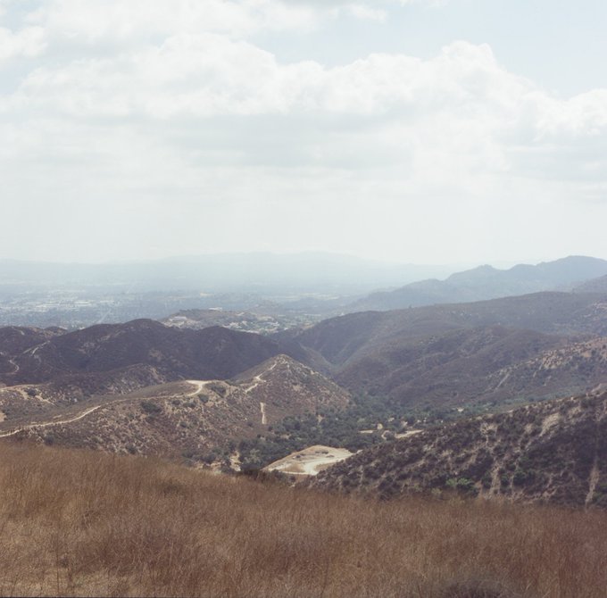 A scenic view from a hillside overlooking dry, rolling mountains with dirt trails and a distant valley city under a hazy, cloudy sky.
