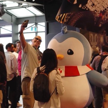 A man in sunglasses takes a selfie with a woman wearing a backpack next to a large mascot costume of Tux, the Linux penguin, at a crowded indoor event.