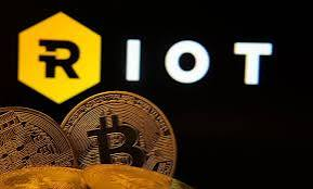 A promotional image showing the RIOT logo (ticker: RIOT) with physical Bitcoin coins displayed in front of it against a dark background.