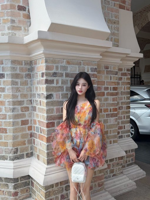A young woman with long dark hair stands against a brick pillar, wearing a colorful floral off-the-shoulder dress and holding a small white quilted handbag.
