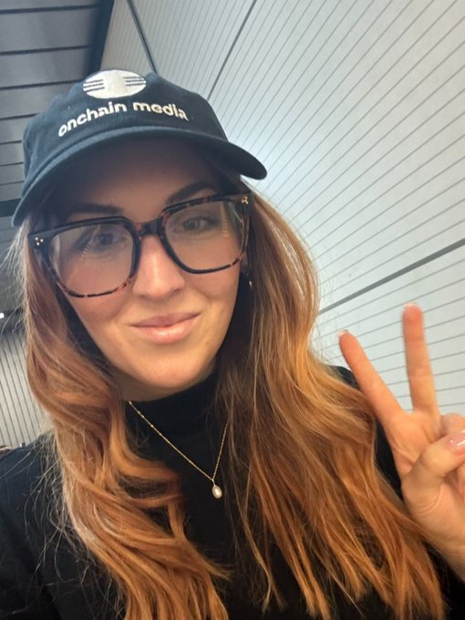 A young woman wearing glasses and a dark baseball cap with 'onchain media' written on it smiles and holds up a peace sign.