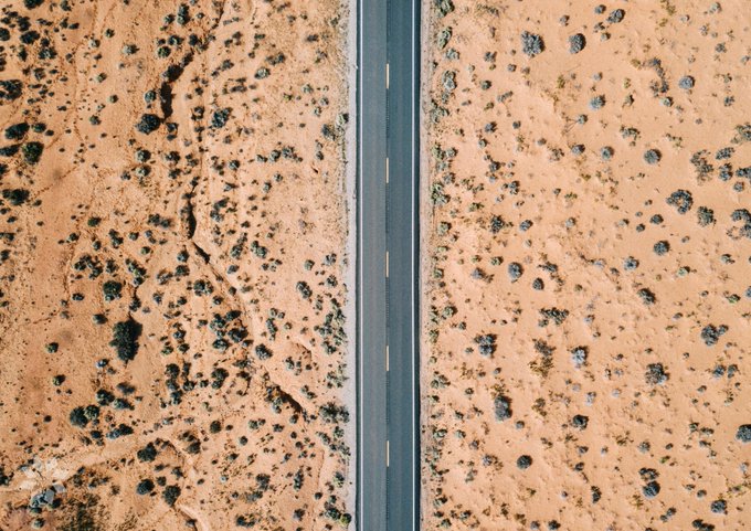 An aerial drone view looking straight down at a straight two-lane paved road with yellow dashes cutting through an arid desert landscape dotted with scrub brush.