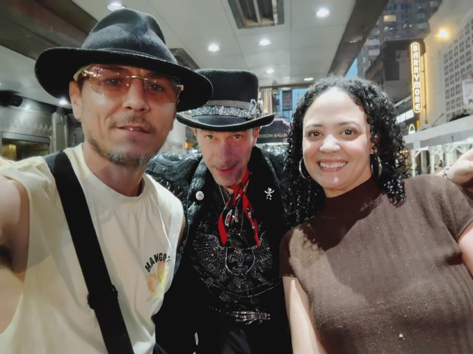 Three people pose for a selfie at night on a city street: a man in a black fedora and suspenders, a man in the center wearing an ornate black cowboy hat and elaborate western-style outfit with red bandana, and a smiling woman with curly hair in a brown top, with a Burger King sign visible in the background.