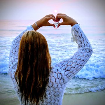 A woman with long red hair, seen from behind, wearing a white knit sweater and forming a heart shape with her hands against a sunset beach background.