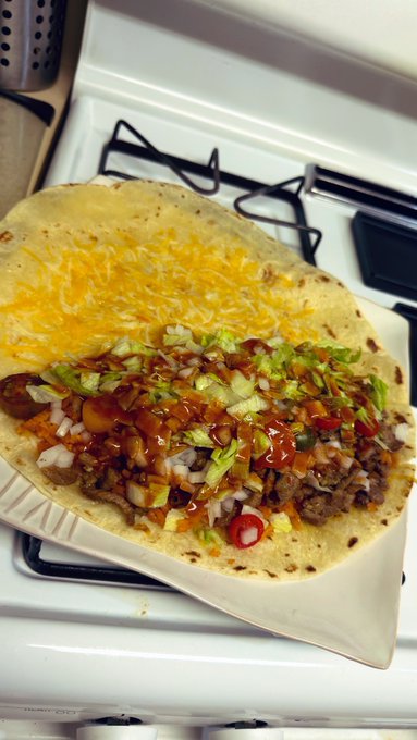 An open-faced flour tortilla topped with shredded cheese, chopped vegetables, ground meat, and hot sauce, resting on a plate atop a stove.