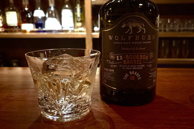 A bottle of Wolfburn Ex-Bourbon Barrel Single Malt Scotch Whisky sits on a wooden bar next to a crystal glass filled with ice.