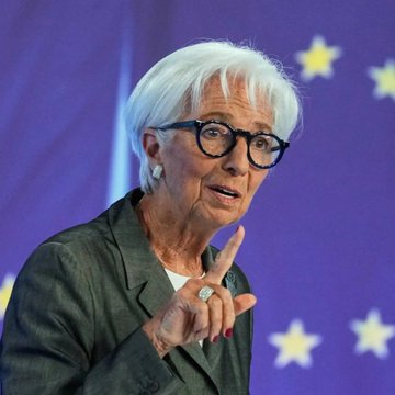 An older woman with short white hair and glasses speaks while pointing her finger, with a European Union flag visible in the background.