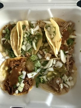Four street-style tacos with carnitas, diced onions, cilantro, and salsa verde served in a foam takeout container, fitting the "$1" taco deal referenced in the caption.
