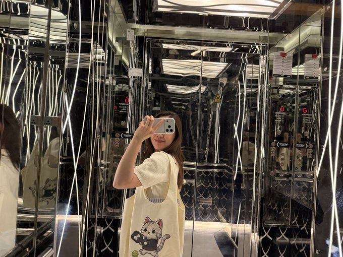A person with long hair, wearing a white shirt and carrying a cat-themed tote bag, takes a mirror selfie with a smartphone inside a mirrored, geometric elevator.