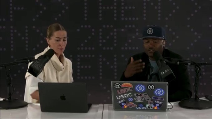 Two people sit at a podcast table with microphones and laptops; the laptop on the right is covered in stickers including 'USDC' logos.