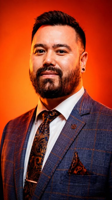 A man with a beard poses for a portrait against an orange background, wearing a blue plaid suit, white shirt, and a paisley tie with matching pocket square.