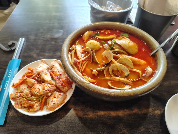 A bowl of spicy Korean fish soup alongside a plate of kimchi sits on a wooden table, paired with chopsticks in a teal wrapper reading "SOUL SOUP." The warm homemade-looking meal illustrates the post's point about enjoying life and treating oneself rather than hoarding crypto gains.