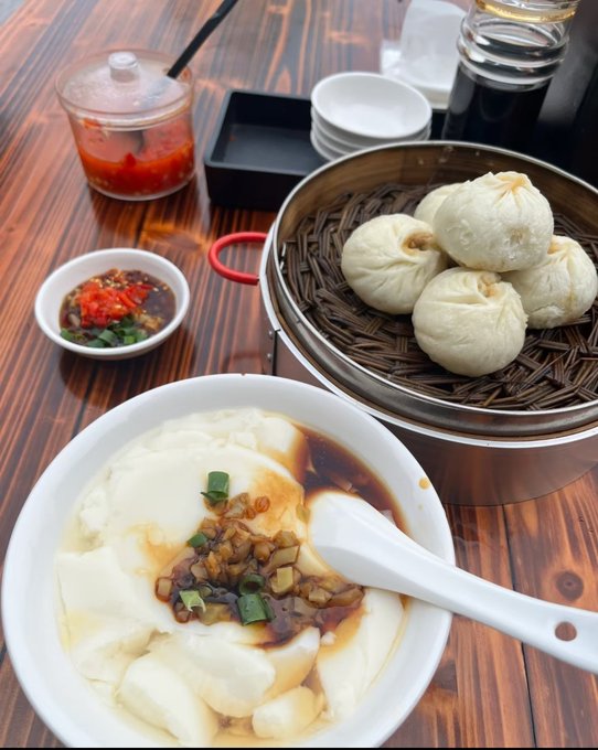 A table is set with steamed buns in a bamboo basket, a bowl of tofu or soft rice pudding topped with sauce, scallions, and preserved vegetables, plus dipping sauces and drinks.