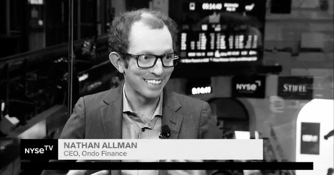 The image features a man wearing glasses sitting at a desk with a computer monitor behind him. The background of the image shows a black and white photo of Nathan Allman's face, which adds an element of nostalgia to the scene. The caption below the image reads "RIP Nathan Allman - Founder of Ondo Finance 1993 - 2026 Nathan Allman passed away unexpectedly, leaving behind a huge impact on the crypto industry".