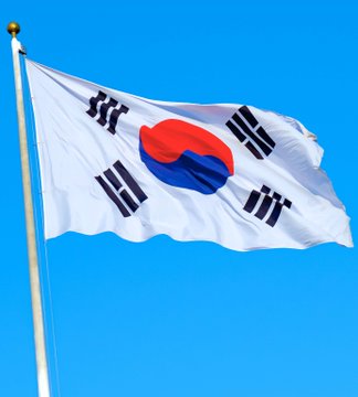 The South Korean flag waves against a clear blue sky, featuring the iconic red and blue taegeuk symbol surrounded by four black trigrams. The image visually represents South Korea as the country where Jeonbuk Bank is launching pioneering bitcoin-backed loans denominated in the local won currency.