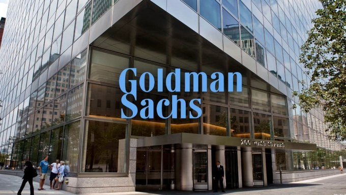 The image shows the exterior of a Goldman Sachs building with the company's name displayed in large blue letters on a glass facade, while a security guard stands at the entrance. The accompanying post highlights Goldman Sachs CEO's public support for U.S. cryptocurrency market structure legislation, visually connecting the financial institution to the breaking news about its stance on crypto regulation.