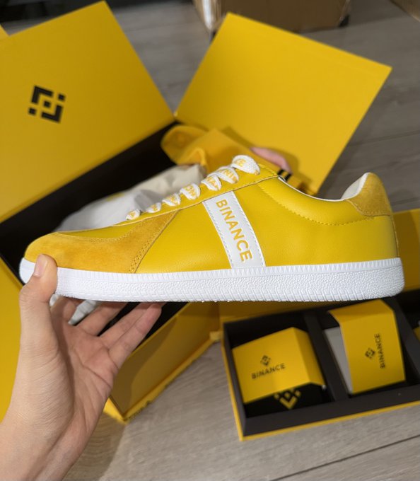 A hand holding up a yellow and white Binance-branded sneaker above its yellow shoebox packaging.