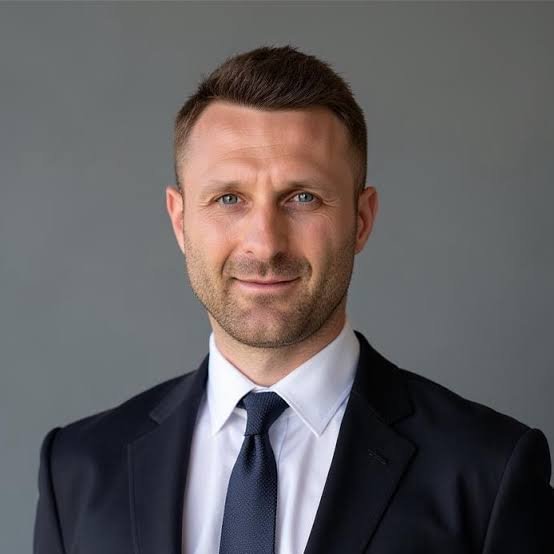 A professional headshot portrait of a man with short brown hair and a beard, wearing a dark suit and tie against a grey background.
