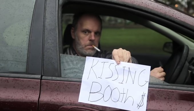 A bearded man sits in a maroon car with a serious expression, smoking a cigarette, while holding up a handwritten sign through the window that reads "KISSING Booth $" — humorously personifying Hunter Biden opening a kissing booth following his laptop scandal, as referenced in the caption.