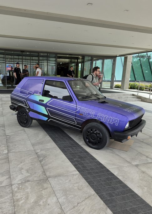 A small blue and purple hatchback car (a Yugo) sits outside a building, wrapped with "SOLANA" branding and angular geometric graphics, mimicking the look of a luxury sports car. The image humorously plays on the post "Lambo? Yugo" by transforming an inexpensive, modest vehicle into a crypto-branded showpiece.
