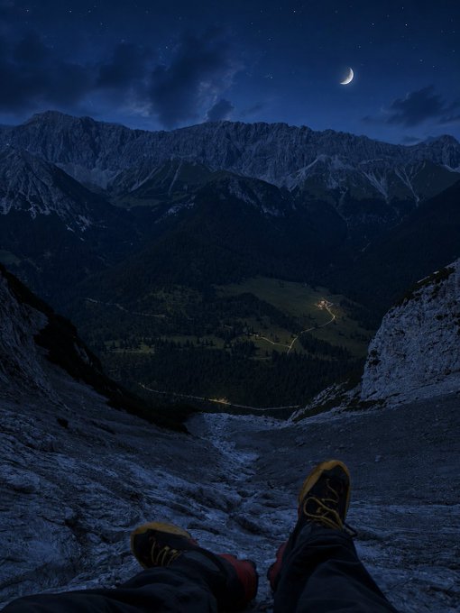 A hiker dangles their booted feet from a rocky cliff edge at night, overlooking a valley dotted with a winding road's headlights beneath a crescent moon and starry sky. The serene, reflective "good night" scene mirrors the post's contemplative close to a thoughtful day of analyzing Ethra Shipping's operational history.