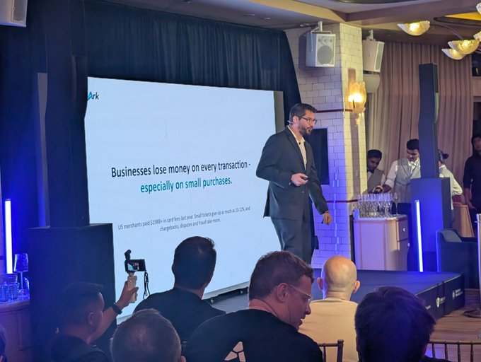 A man in a suit presenting at a business event. Slide reads: 'Businesses lose money on every transaction - especially on small purchases.' Subtext mentions US merchants paid $338B+ last year in card fees.