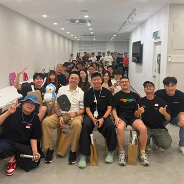 A group photo of approximately fifteen smiling people posing together in a brightly lit hallway, with some individuals in the front row holding plush toys and wearing black t-shirts.