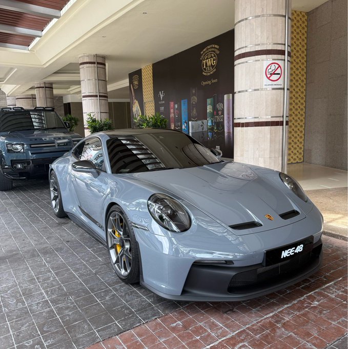 The image features a silver Porsche 911 sports car parked outside, possibly at a showroom or dealership. The car has a black roof and white wheels, giving it an elegant appearance. In front of the car, there are two potted plants adding a touch of greenery to the scene. The background includes a building with columns and a sign that reads "NEE 44". The image also contains text that says "@KASTxyz", which could be related to the Porsche 911 sports car or another product associated with it.
