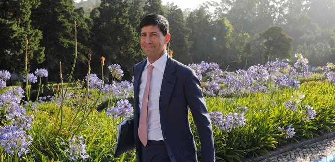 A man in a navy suit and pink tie stands smiling amid a garden of purple agapanthus flowers, fittingly mirroring the post's news of US CPI inflation holding steady at an expected 3.4%. The tranquil outdoor backdrop underscores the report's steady, in-line nature—neither alarming nor celebratory.