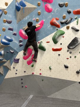 A person climbing an indoor bouldering wall, reaching up to grip a large pink hold among various colorful climbing grips.