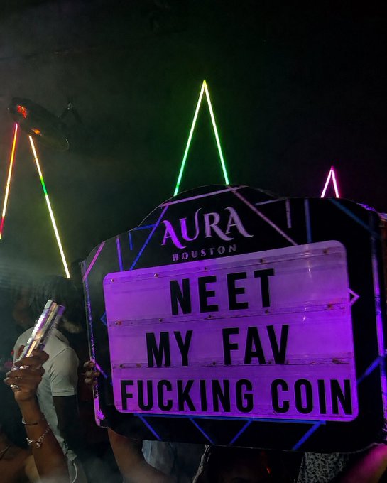 The image shows a person holding up a sign that reads "Neat my fav crap coin", which seems to be promoting a product or service. There are other people present, but they are not the main focus of the scene. The background features neon lights, suggesting that this might be taking place in an event space like a club or a bar.