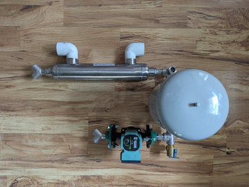 An industrial assembly of stainless steel piping components including a heat exchanger or UV sterilizer, valves, and a white pressure tank, laid out on a wooden floor.