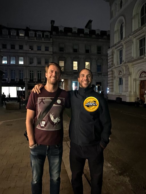 Two men pose together at night in front of a grand historic building, one wearing a maroon graphic t-shirt and the other in a black jacket featuring a yellow "SPX 6900" patch, with the post playfully referencing the SPX6900 meme coin and a joke about being mistaken for Dr. Strange or "6/7."