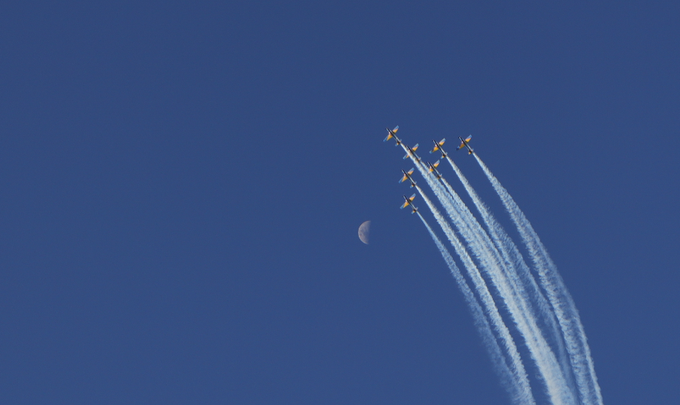 A formation of jets flies through a clear blue sky, leaving white contrails behind them, with the moon visible to the left.