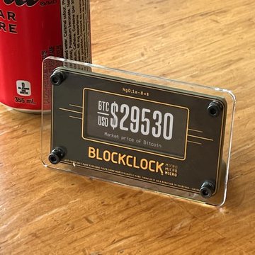 A Blockclock device displays a live Bitcoin price of BTC/USD $29,530 on a wooden surface next to a beverage can. The image satirizes the post by suggesting this real-time price tracker could double as an exploit tool for locating "pleb nodes," mocking overly paranoid crypto conspiracy theories.