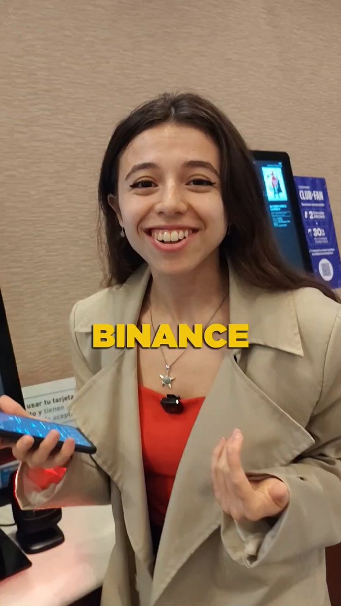 The image features a woman wearing a tan jacket, holding a cell phone with a blue case. She has her hand outstretched as if she's about to make a payment or share something interesting on social media. The background of the photo shows a computer monitor and a cash register, suggesting that this scene takes place in an office setting. The caption "Binance Pay in Argentina" is written above the woman, which might be related to her location or the context of the image.