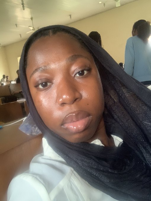 A selfie of a young woman wearing a dark hijab and white top, taken indoors in what appears to be a classroom setting.