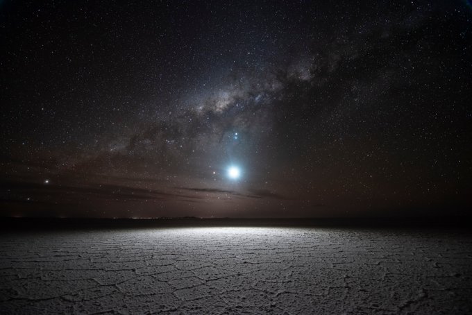 A night sky photograph showing the Milky Way galaxy stretching across the sky, with a bright celestial object (possibly a planet) shining over a cracked, dry salt flat landscape.