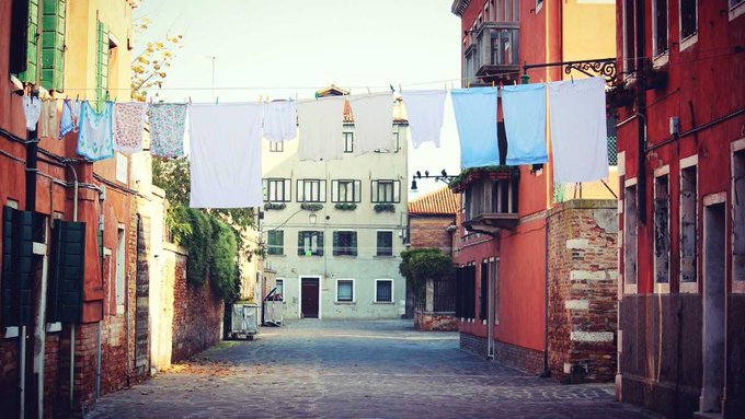A narrow European street scene with white and blue laundry hanging on a clothesline strung between warm-toned, weathered buildings.