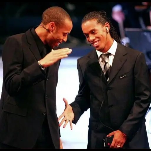 Two men in dark suits share a moment together on stage, smiling and gesturing toward each other, one holding a microphone.