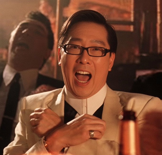 An Asian man wearing glasses and a beige suit excitedly shouts with clenched fists at a celebratory gathering, with another man laughing behind him.