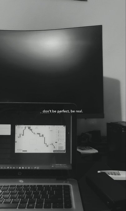 A black-and-white photo shows a laptop in front of a curved monitor displaying the quote 'don't be perfect, be real,' with a stock chart visible on the laptop screen.